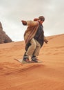 Young woman posing as sand dune surfing wearing bisht - traditional Bedouin coat. Sandsurfing is one of the attractions in Wadi
