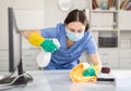 Young woman in a protective mask, cleaning the office, wipes the desktop
