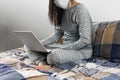 Young woman in a protective mask is sitting in the bed and using her laptop. Home office concept. Royalty Free Stock Photo