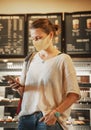 A young woman in a protective mask stands in a cafe at the counter with desserts and a cash register with a smartphone in her Royalty Free Stock Photo