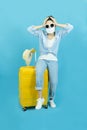 A young woman with a protective medical mask on her face, sitting on a yellow suitcase.The concept of travel, quarantine and