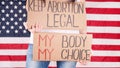Young woman protester holds cardboard with Keep Abortion Legal and My Body My Choice signs against USA flag on background. Girl Royalty Free Stock Photo