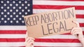 Young woman protester holds cardboard with Keep Abortion Legal sign against USA flag on background. Girl protesting against anti-