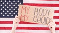 Young woman protester holds cardboard with My Body My Choice sign against USA flag on background. Girl protesting against anti-