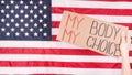 Young woman protester holds cardboard with My Body My Choice sign against USA flag on background. Girl protesting against anti-