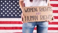 Young woman protester holds cardboard with Womens Rights Are Human Rights sign against USA flag on background. Feminist power.