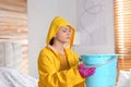 Young woman in raincoat collecting leaking water from ceiling at home. Time to call roof service Royalty Free Stock Photo