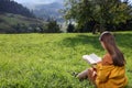 Young woman reading book on green meadow in mountains, back view Royalty Free Stock Photo