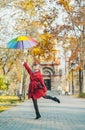 Young woman in red coat with a rainbow colorful umbrella jumping over autumn walkway in city park Royalty Free Stock Photo
