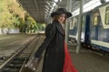 Young woman in red dress, black retro coat and hat holds vintage suitcase and stands on platform near train