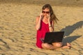 Young woman in red dress with computer and smartphone on the beach. Freelance and downshifting concept.