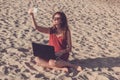 Young woman in red dress with computer and smartphone on the beach. Freelance and downshifting concept.