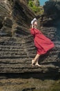 A young woman in a red dress and white hat stands on a rock by the sea and holds a hat with her hands. The face is illuminated by Royalty Free Stock Photo