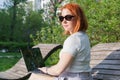 Young woman with red hair with a laptop on a park bench. Freelancer woman works in the park