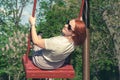 Young woman with red hair in sunglasses smiles swinging on a swing in a city park. Childrens entertainment for adults Royalty Free Stock Photo