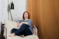 Young woman with red hair wearing jeans and knitted pullover sitting on cozy armchair in interior of loft