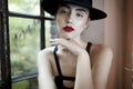 Cheerful young woman with red lips, in black hat, sitting on chair, touching her chin with hand, behind a window. Royalty Free Stock Photo