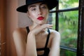 Young woman with red lips, in black hat, sitting on chair, touching her chin delicate with hand, behind a window. Royalty Free Stock Photo