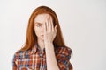 Young woman with red long hair and pale skin, covering half of face and looking serious at camera, standing over white Royalty Free Stock Photo