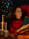 Young woman in a red pyjama reads a book by the burning candles and glowing Christmas tree with festive decorations