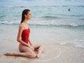 A young woman in a red swimsuit sits on the sand with a beautiful sun tan and looks out at the ocean in the tropics on Royalty Free Stock Photo