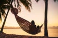 Young woman relaxing in hammock hinged between palm trees on the sand beach at orange sunrise morning time Royalty Free Stock Photo