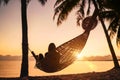 Young woman relaxing in hammock hinged between palm trees on the sand beach at orange sunrise morning time