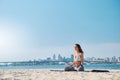 Young woman relaxing practicing yoga and meditation on the sand beach with city background. Sporty girl is sitting in Royalty Free Stock Photo