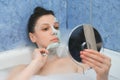 Young woman is removing alginate mask from her face lying in bathroom with foam.