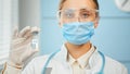 Young woman researcher in blue disposable mask glasses and white coat holds transparent vial with vaccine