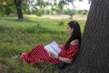 Young woman is resting on a summer day in the park. A brunette with a book on the grass near a tree sits with her back to the