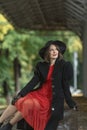 Young woman in retro clothes at train station. Aristocratic woman wears red dress, black coat and wide-brimmed hat