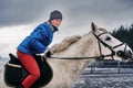 Young woman rider in a blue blazer and sporting a cap for a walk on a white horse on a cloudy winter day. Royalty Free Stock Photo