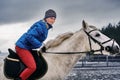 Young woman rider in a blue blazer and sporting a cap for a walk on a white horse on a cloudy winter day.