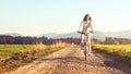 Young woman rides bicycle on dusty country road, afternoon sun shines at her, view from front Royalty Free Stock Photo