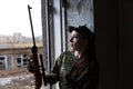 A young woman with a rifle in uniform at the window looking at the street. The woman sniper in a green suit and cap. Royalty Free Stock Photo