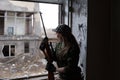 A young woman with a rifle in uniform at the window looking at the street. The woman sniper in a green suit and cap