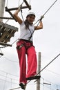 A young woman in a rope amusement park walks a tightrope in a safety harness and a helmet