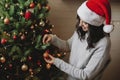 Young woman in santa hat decorating modern christmas tree with glitter bauble in festive room