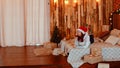 Young woman in santa hat and glasses lying on bed and drinking red wine. Pretty female resting with alcohol on holiday