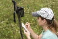 Young woman scientist zoologist writing down data from trap camera to notepad, observing wild animals in taiga forest, monitoring