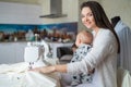 A young woman sews in the kitchen and holds a small child. Mom teaches her little son to sew on a sewing machine. Self Royalty Free Stock Photo