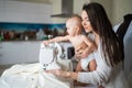 A young woman sews in the kitchen and holds a small child. Mom teaches her little son to sew on a sewing machine. Self Royalty Free Stock Photo