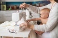 A young woman sews in the kitchen and holds a small child. Mom teaches her little son to sew on a sewing machine. Self Royalty Free Stock Photo