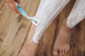 Young woman shaving legs with plastic razor and depilation cream in home bathroom with green plants. Skin care. Hand holding Royalty Free Stock Photo