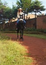 Young woman in shirt and straw hat, riding black horse in the park over dirt road, brick wall near her, on sunny morning