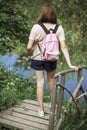 A young woman with short hair and a pink backpack stands with her back to the camera on a small wooden bridge near a river in the Royalty Free Stock Photo