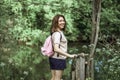 A young woman with short hair and a pink backpack stands on a small wooden bridge near a river in the woods, looking at the camera Royalty Free Stock Photo