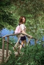 A young woman with short hair and a pink backpack stands on a small wooden bridge near a river in the woods, looking at the camera Royalty Free Stock Photo