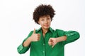 Young woman shows thumbs up and thumbs down with doubt, confused, making her choice, standing over white background Royalty Free Stock Photo